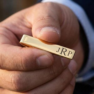 May include: A gold-colored tie bar with the engraved initials "JRP" in a classic font. The rectangular bar is held in a person's hand, showcasing its polished, reflective surface. This accessory is a stylish addition to any formal attire.