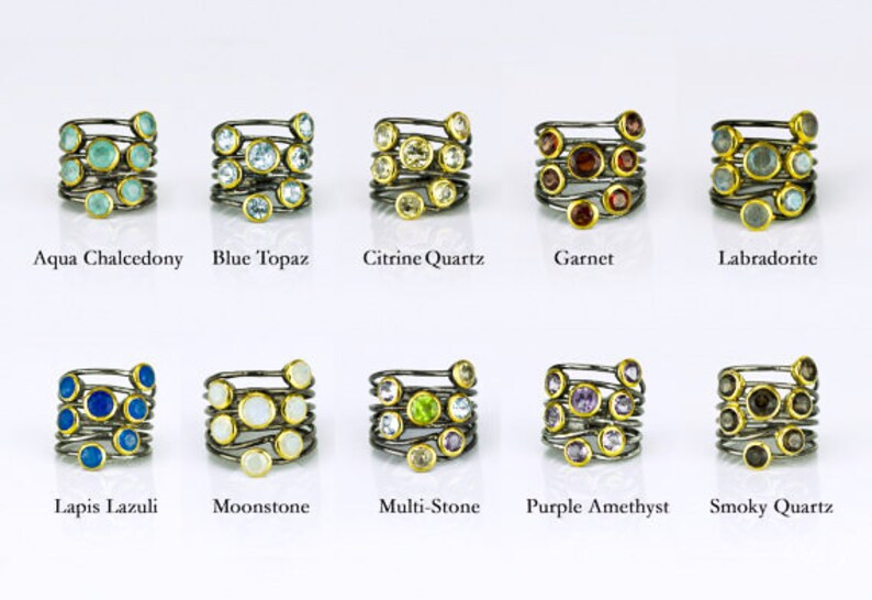 May include: Ten silver rings with gold accents, each featuring a different gemstone. The gemstones are arranged in a spiral pattern on each ring. The gemstones include aqua chalcedony, blue topaz, citrine quartz, garnet, labradorite, lapis lazuli, moonstone, multi-stone, purple amethyst, and smoky quartz.