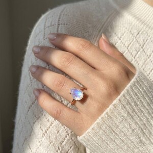 Moonstone teardrop ring worn on hand with cozy cream sweater, showing the delicate prong setting and blue flash of the stone. A June birthstone ring available in sizes 5 to 9, with half sizes upon request, ideal as a gift for her.