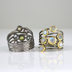 May include: Two silver rings with multiple bands and colorful gemstones. The ring on the left has a variety of small gemstones in shades of blue, green, purple, and white. The ring on the right has larger, round, iridescent gemstones.