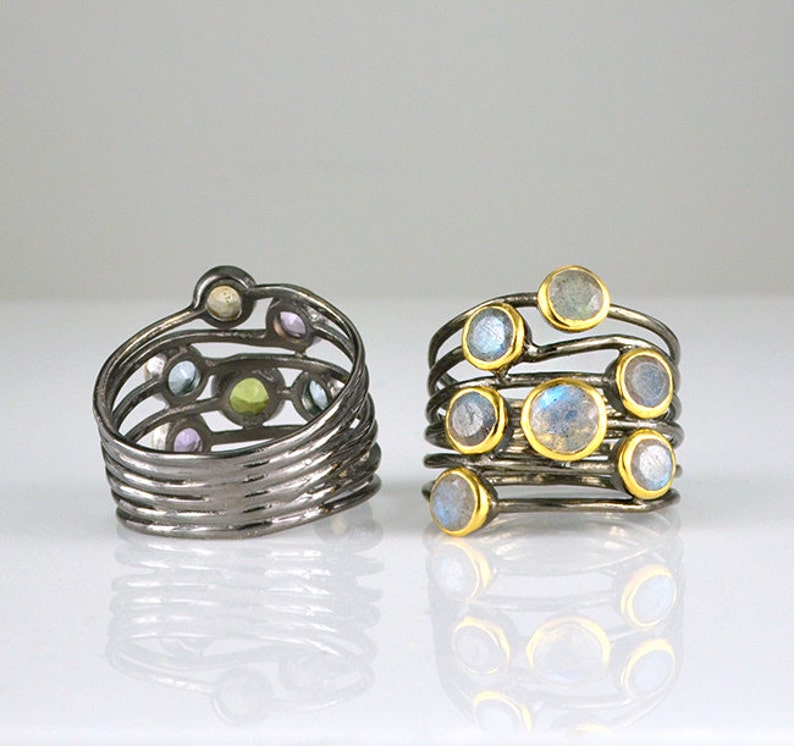 May include: Two silver rings with multiple bands and colorful gemstones. The ring on the left has a variety of small gemstones in shades of blue, green, purple, and white. The ring on the right has larger, round, iridescent gemstones.