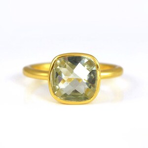 May include: A gold ring with a light green gemstone. The gemstone is a cushion cut and is set in a bezel setting.
