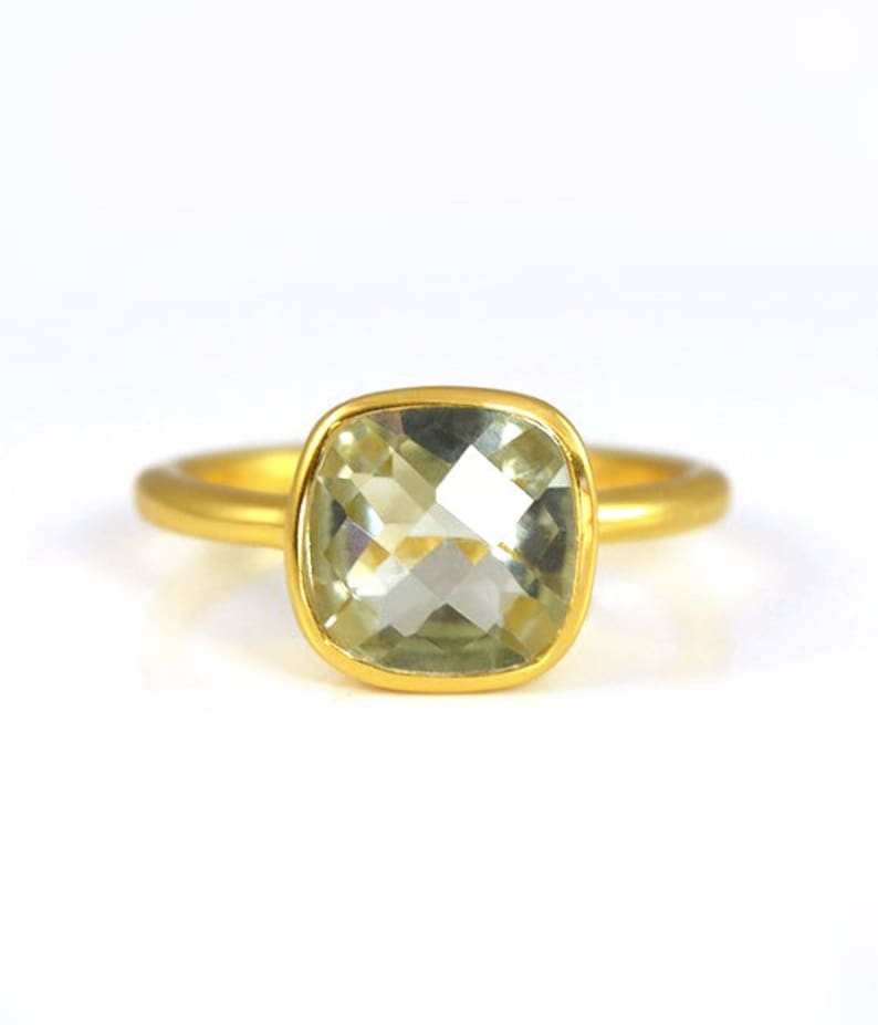 May include: A gold ring with a light green gemstone. The gemstone is a cushion cut and is set in a bezel setting.