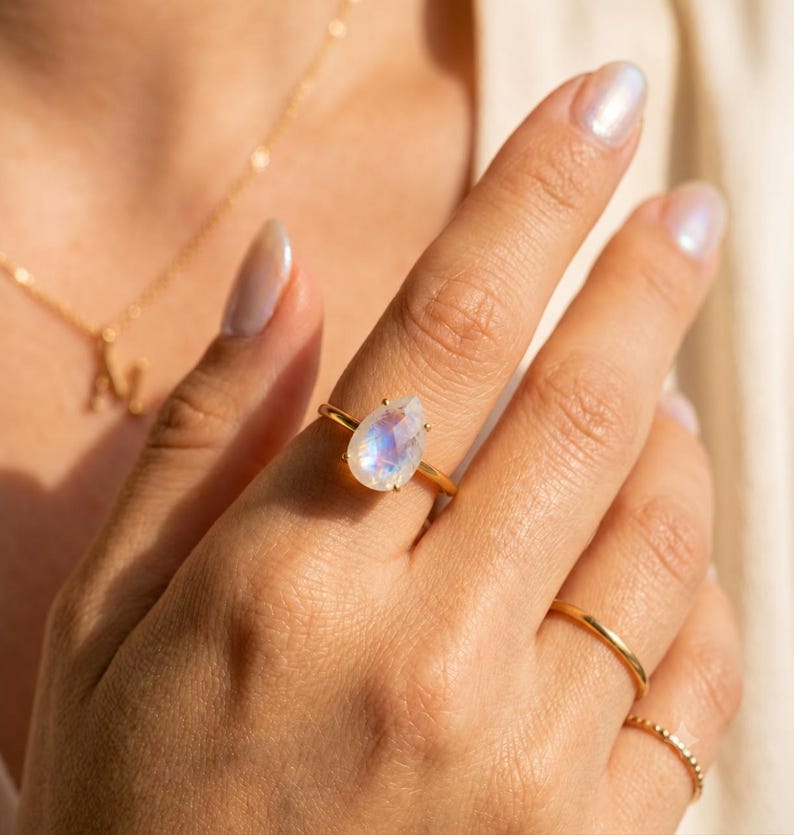 Moonstone teardrop ring worn on hand in warm sunlight, featuring a pear-shaped rainbow moonstone with blue flash in a prong setting on a gold band. A June birthstone ring, gemstone statement ring, gift for her, birthday gift, anniversary gift, or meaningful everyday jewelry piece.