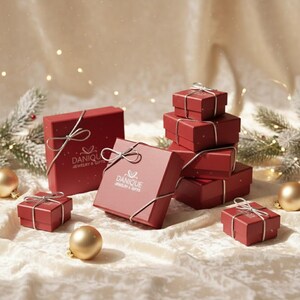 Red Danique Jewelry gift boxes tied with silver ribbon on ivory satin, surrounded by gold ornaments and evergreen sprigs. Holiday packaging photo showing elegant, ready-to-gift personalized jewelry for Christmas, anniversary, or Mother’s Day.