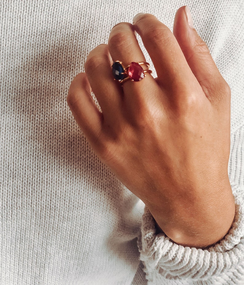 May include: A hand wearing three gold rings with red and blue gemstones. The rings are stacked on the middle finger.