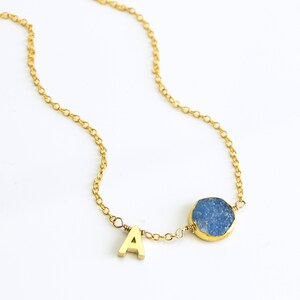 K&ouml;nnte beinhalten: Eine goldene Kettenkette mit einem Anh&auml;nger aus blauem Achat und einem goldenen Buchstaben 'A' als Anh&auml;nger.