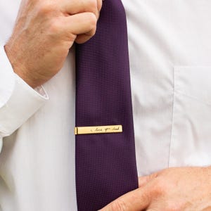 May include: A gold-colored tie bar with the handwritten inscription "i love you dad" is clipped to a dark purple tie. The tie is worn with a white dress shirt.