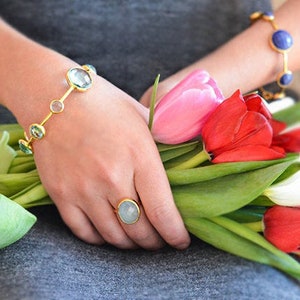 May include: A gold bracelet with oval-shaped light blue gemstones and a gold ring with a light blue oval gemstone. The jewellery is worn by a person holding a bouquet of pink, red and white tulips.