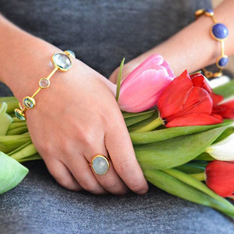 May include: A gold bracelet with oval-shaped light blue gemstones and a gold ring with a light blue oval gemstone. The jewellery is worn by a person holding a bouquet of pink, red and white tulips.