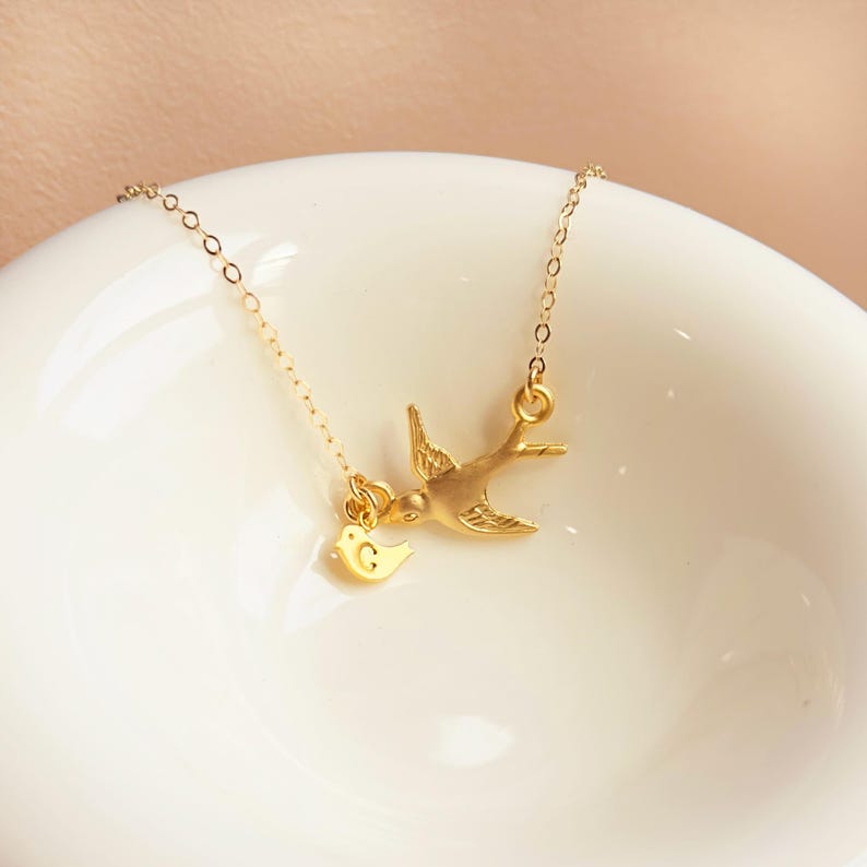 Gold mama bird necklace photographed on a white plate, delicate dainty jewelry flatlay, personalized initial charms for children, modern family necklace.