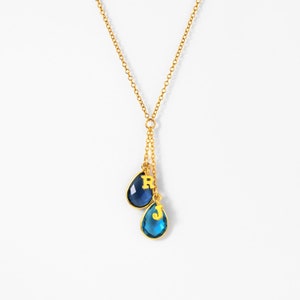 May include: A gold chain necklace with two teardrop-shaped blue gemstone pendants. Each pendant has a gold letter charm, one with the letter 'R' and the other with the letter 'J'.