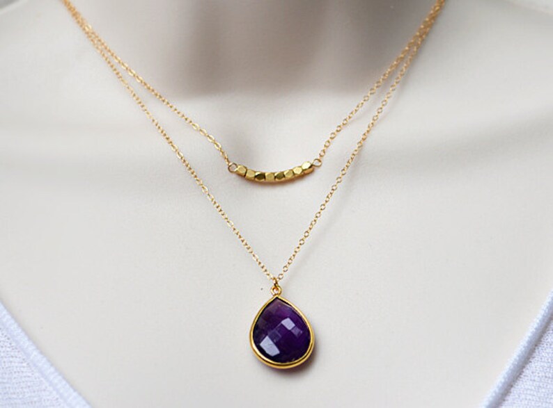 Pode incluir: Um colar de corrente de ouro com um pingente de pedra preciosa de cor p&uacute;rpura em forma de l&aacute;grima e uma corrente de ouro menor com uma fila de contas douradas.