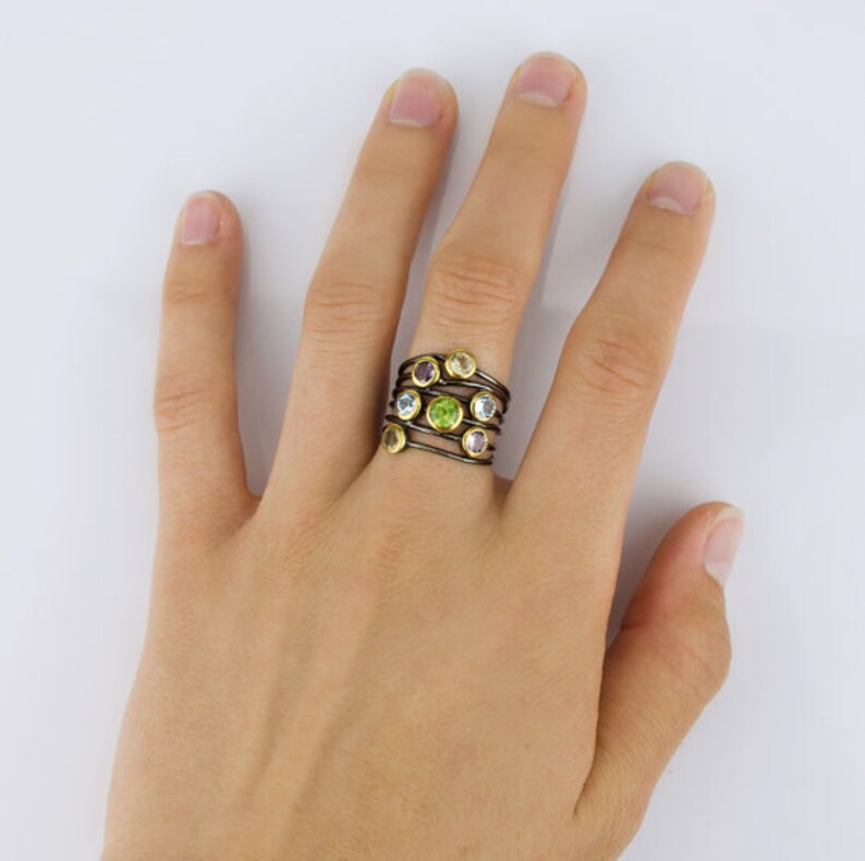 May include: A multi-colored gemstone ring with a black and gold band. The ring features a variety of round gemstones, including green, yellow, purple, and blue.