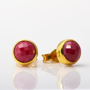 May include: A pair of gold stud earrings with red ruby gemstones. The earrings have a round shape and a smooth, polished finish.