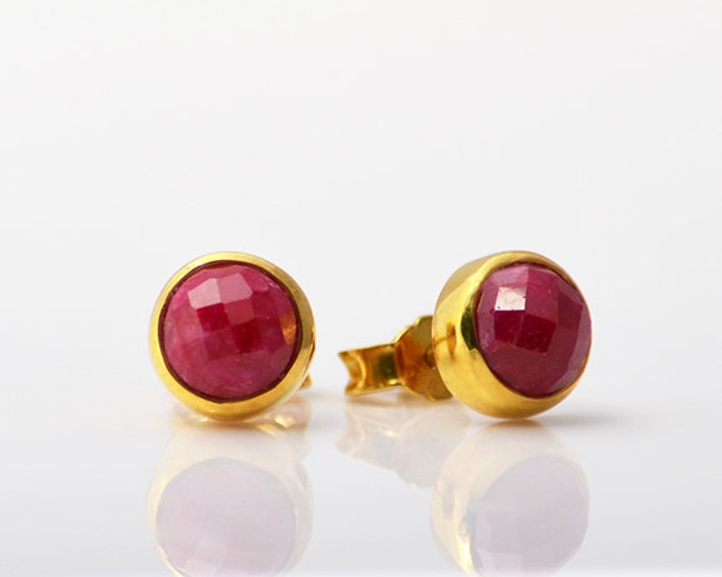 May include: A pair of gold stud earrings with red ruby gemstones. The earrings have a round shape and a smooth, polished finish.