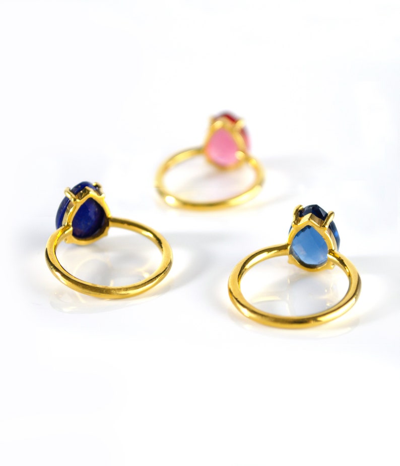 May include: Three gold rings with pear-shaped gemstones. The rings are on a white surface. The gemstones are blue, pink, and light blue.