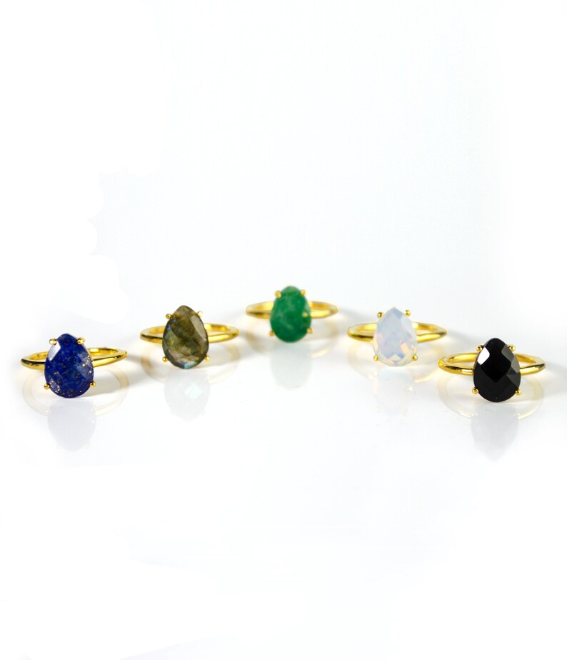 May include: Five gold rings with different colored gemstone settings. The rings feature teardrop-shaped stones in shades of blue, green, white, black, and gray.