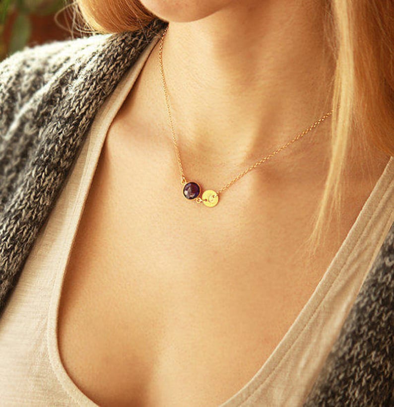 May include: A gold chain necklace with two small round pendants. One pendant is a dark purple gemstone and the other is a gold disc with a small engraved letter.