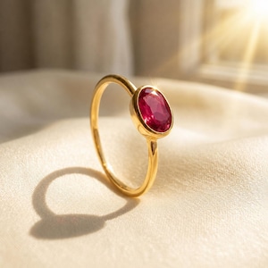 May include: A gold ring featuring a single oval-cut deep pinkish-red gemstone set in a smooth metal bezel. The band is thin and polished, showcasing a bright metallic luster that reflects light across the surface of the setting and the stone.