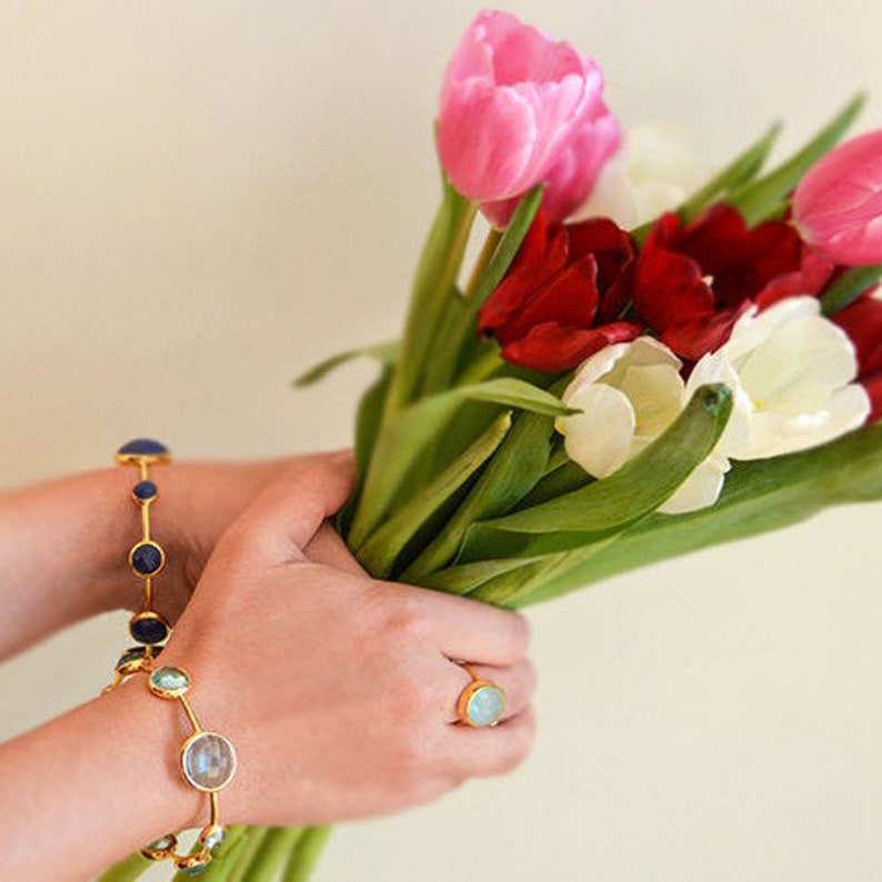 May include: A pair of gold bracelets with blue and green gemstones. The bracelets are worn on a person's wrist, who is holding a bouquet of pink, red and white tulips.