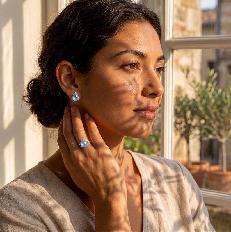 Lifestyle image of model wearing moonstone ring and moonstone stud earrings near a window, showing coordinated birthstone jewelry in warm natural light. A June birthstone ring, rainbow moonstone jewelry set, bridesmaid gift, or birthday gift for her.