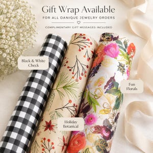 May include: Three rolls of gift wrap featuring distinct patterns: a black and white checkered print, a tan paper with red and green botanical illustrations, and a white paper with colorful floral designs. Text reads Gift Wrap Available.