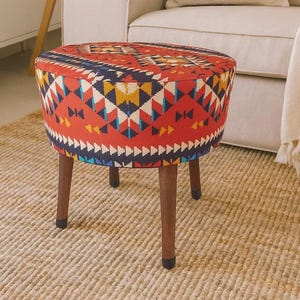 May include: A round ottoman featuring a vibrant red fabric upholstery with a multi-colored geometric pattern in shades of blue, yellow, white, and black. The piece is supported by four dark brown wooden legs that taper toward the floor.