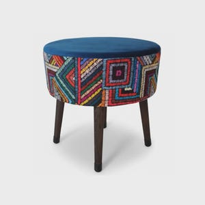 May include: A round stool with a solid dark blue velvet top and a base upholstered in a multi-colored fabric featuring a geometric pattern of squares and triangles in red, yellow, blue, and white. The stool stands on four dark brown wooden legs.