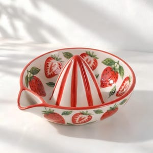 May include: A white ceramic citrus juicer featuring a central reamer with red vertical stripes. The bowl is decorated with hand-painted red strawberries and green leaves, and includes a small pouring spout on the left side of the rim.