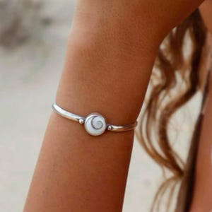 May include: A silver-toned metal bangle bracelet featuring a circular centrepiece with a white and grey spiral shell inlay. The bracelet has a smooth, polished finish and a simple, clean design that sits securely around the wrist area.