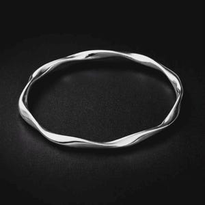 May include: A polished silver bangle bracelet featuring a continuous, fluid, and undulating wave-like design. The metallic surface reflects light, highlighting the smooth, curved contours of the circular band against a dark, neutral background.
