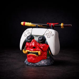 May include: A white video game controller stand featuring a red and gold oni mask base with a sculpted texture. A miniature black, gold, and red katana sword rests horizontally across the top of the controller, which is held in place by the stand.