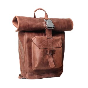 Pode incluir: Uma mochila de couro castanho com fecho de enrolar, apresentando um bolso frontal grande com aba e um mecanismo de fivela de metal prateado para prender o compartimento principal. A bolsa inclui uma alça superior e costuras visíveis.