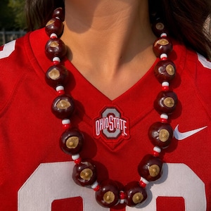 May include: A necklace composed of dark brown, polished buckeye nuts alternating with small, round white and red beads. The necklace is worn around the neck, resting against a red athletic jersey featuring the Ohio State logo and white numbers.