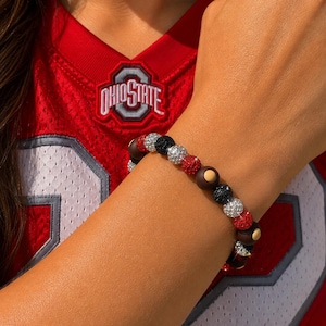 Könnte beinhalten: Ein Perlenarmband mit einem wiederkehrenden Muster aus runden, texturierten, glitzernden Perlen in Rot, Silber und Schwarz, durchsetzt mit glatten, matten braunen Perlen, die einen kleinen, zentrierten beigen Punkt aufweisen.