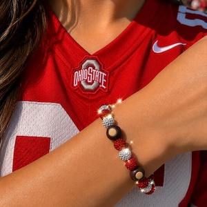 Könnte beinhalten: Ein Perlenarmband mit einem sich wiederholenden Muster aus roten, silbernen und dunkelbraunen kugelförmigen Perlen. Die roten und silbernen Perlen haben eine texturierte, glitzernde Oberfläche, während die dunkelbraunen Perlen glatt sind und einen kleinen, hellen kreisförmigen Mittelpunkt aufweisen.