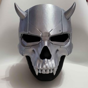 May include: A metallic silver skull mask featuring two prominent curved horns on the upper sides, deep eye sockets, a nasal cavity, and a set of sharp, pointed teeth. The design includes visible panel lines and mechanical details across the surface.