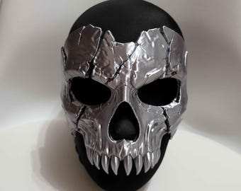 May include: A metallic silver skull mask featuring a cracked, fragmented surface texture. The mask has deep, hollow eye sockets and a prominent nasal cavity, with sharp, pointed teeth along the jawline, all set against a solid black base.