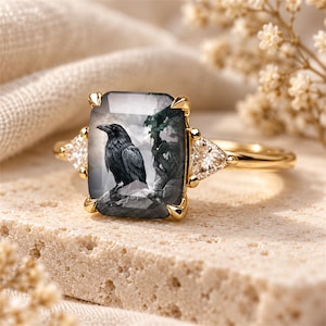 May include: A gold ring featuring a large rectangular center stone with a detailed black and white illustration of a raven perched on a branch. The center stone is flanked by two smaller triangular clear stones set in a polished gold band.