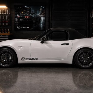 Op de afbeelding: Een witte Mazda MX-5 Miata cabriolet met een zwarte softtop en donkergrijze meerspaaks wielen. Het voertuig is voorzien van een zwarte Mazda-logosticker op het onderste zijpaneel. De auto staat geparkeerd in een garage met een Mazda-poster.