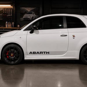 Op de afbeelding: Een witte Abarth 595 hatchback in zijaanzicht, voorzien van zwarte meerspaaks lichtmetalen velgen, rode remklauwen en zwarte Abarth-belettering op het onderste zijpaneel. Het voertuig toont het Abarth-schorpioenembleem op het achterpaneel.