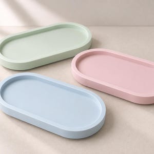 Könnte beinhalten: Drei ovale Tabletts in Pastellfarben, darunter ein hellgrünes, ein hellrosa und ein hellblaues Tablett. Jedes Tablett hat eine glatte, matte Oberfläche mit einem erhöhten Rand und einer flachen, vertieften Mitte, die in einer Gruppe angeordnet sind.
