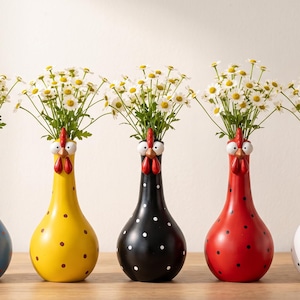 Peut inclure: Un ensemble de cinq vases en céramique en forme de poules, chacun avec une couleur de base différente : bleu, jaune, noir, rouge ou blanc avec des pois noirs ou rouges. Chaque vase présente une tête de poule avec des yeux, un bec et une crête rouge.