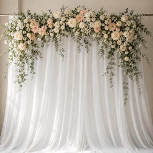 May include: A white sheer fabric backdrop draped in vertical folds, adorned with a large floral arrangement across the top. The arrangement features cream and pale peach roses, dahlias, and small white blossoms interspersed with green eucalyptus foliage.