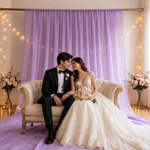 May include: A cream-colored tufted loveseat featuring curved wooden legs, positioned in front of a light purple fabric backdrop draped with warm string lights. Two floral bouquets in glass vases are placed on either side of the seating area.