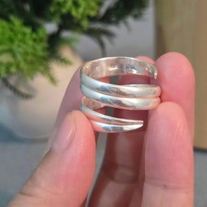 Può includere: Un anello color argento lucido caratterizzato da un design bypass con due fasce sovrapposte che si avvolgono a spirale attorno al dito. Il metallo presenta una superficie liscia e riflettente e le estremità delle fasce sono affusolate.