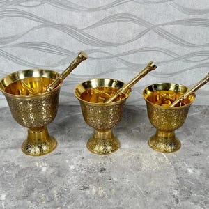 May include: Three gold-coloured brass mortar and pestle sets of varying sizes. Each mortar features a pedestal base and an intricate floral pattern engraved across the exterior surface. Each set includes a matching slender pestle tool.