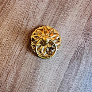 May include: A circular brass drawer pull featuring a raised floral petal design on the backplate and a central swinging ring handle. The metal surface has a polished gold-tone finish with visible texture and two small screw holes on the sides.