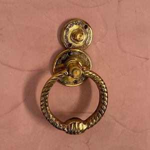 May include: A gold-toned metal door knocker featuring a circular, twisted rope-textured ring attached to a decorative mounting plate. The plate displays an intricate, engraved leaf-like pattern around the central post and screw holes.